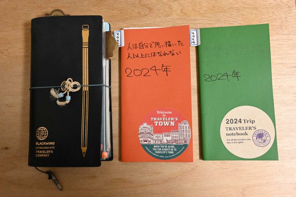 トラベラーズノート × BLACKWINGコラボ。叶結びのアクセサリーをつけている。横には2冊のトラベラーズノートのリフィル。